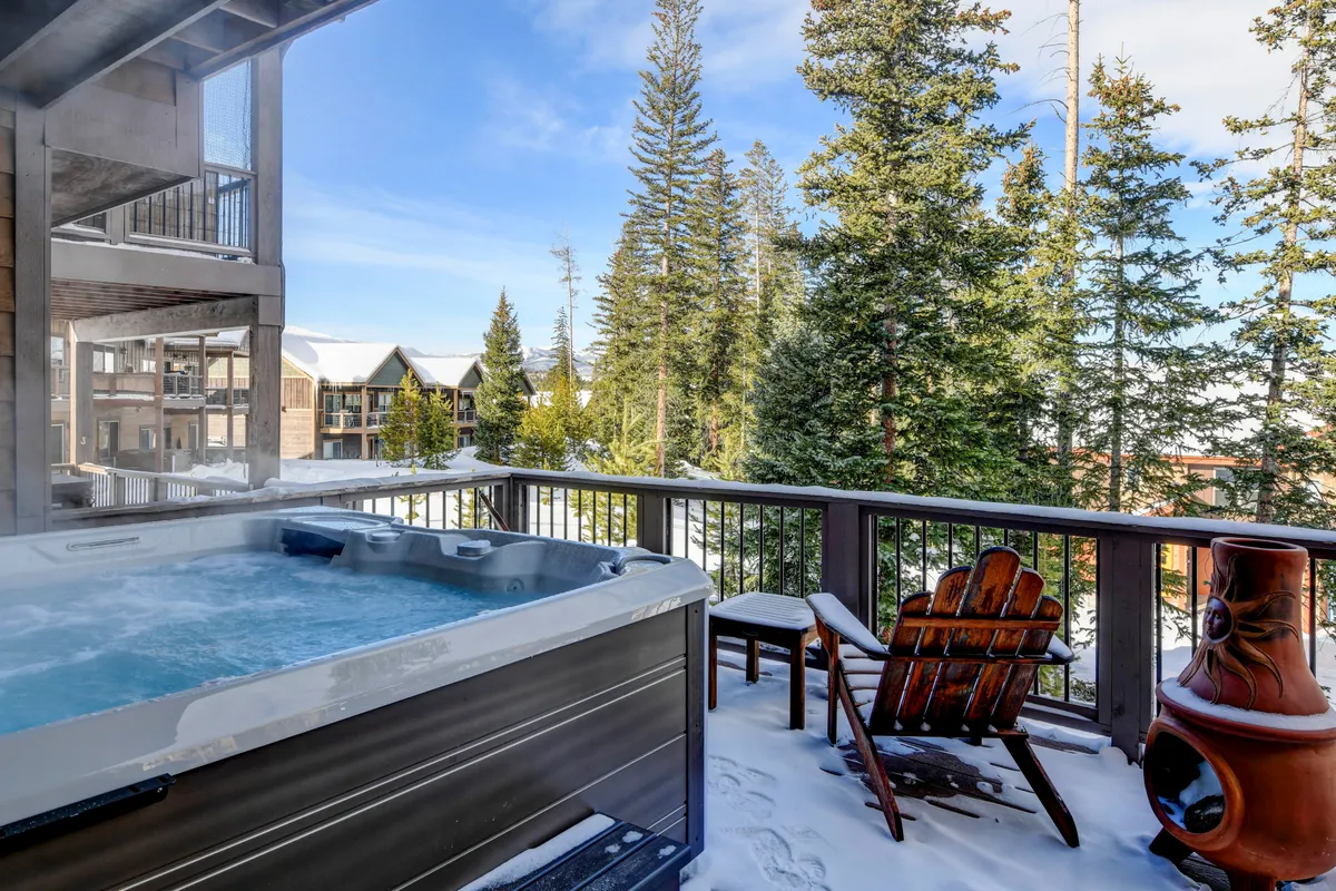 An inviting hot tub sits among tall trees on a snow-covered patio, with wooden chairs nearby. The view suggests peaceful relaxation after a day outdoors—perhaps stargazing or watching the sunset, with the snow adding to the mountain ambiance.
