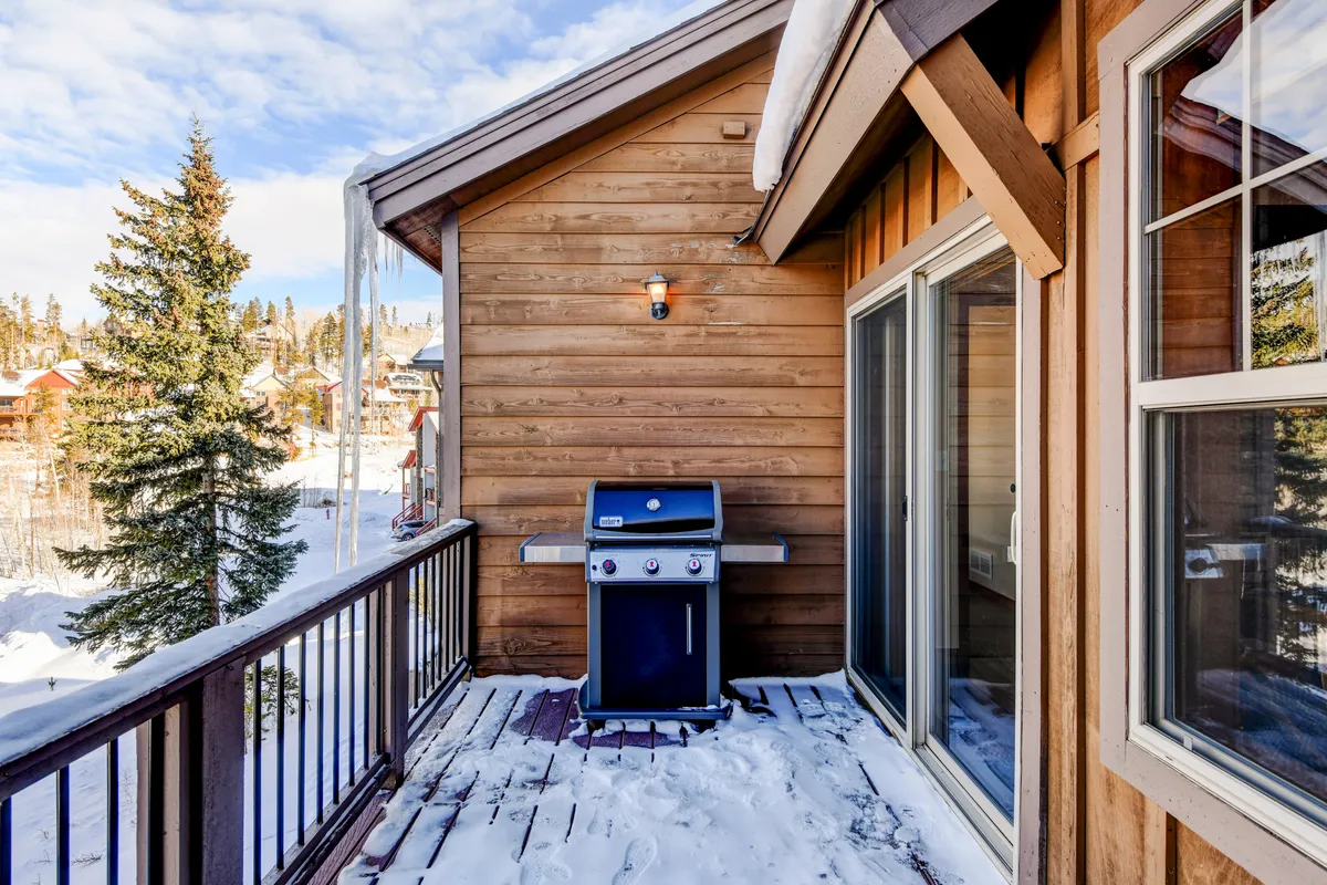An exterior view of a wooden deck with a grill and snow-covered landscape, accented by a pine tree. The space is set up for outdoor dining and barbecuing, offering a scenic mountain backdrop for alfresco meals.