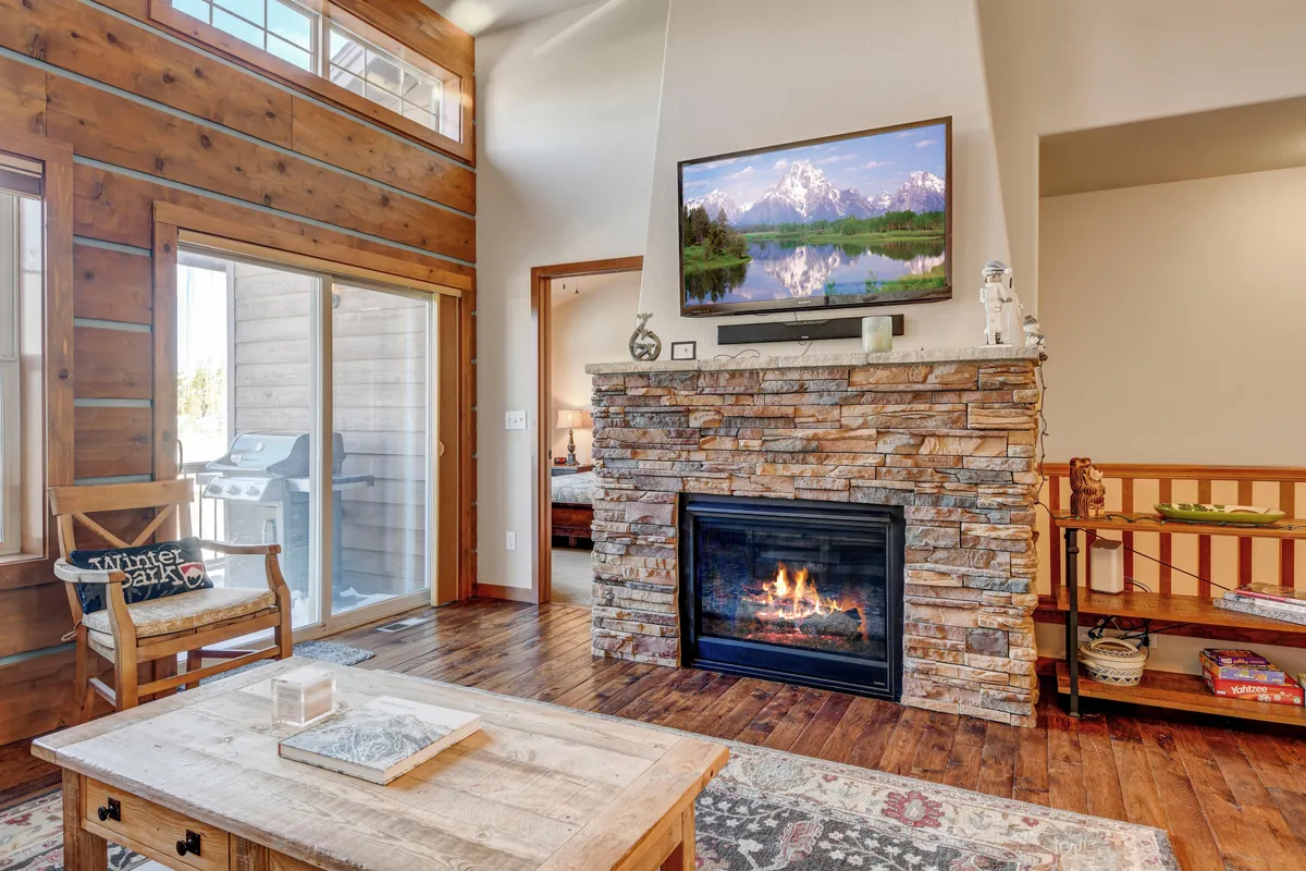 A spacious living room with large floor-to-ceiling windows letting in ample natural light. It features a modern fireplace, a wall-mounted television, wooden beams overhead, and a rustic wooden coffee table. Plush seating is arranged to maximize comfort and the view outside.