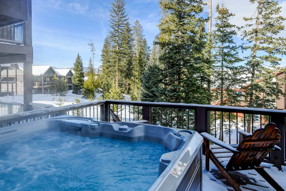 An inviting outdoor hot tub surrounded by snow-covered ground, trees, and a deck railing, with an Adirondack chair nearby. The peaceful setting offers views of nature, suggesting relaxation after a day of adventure.