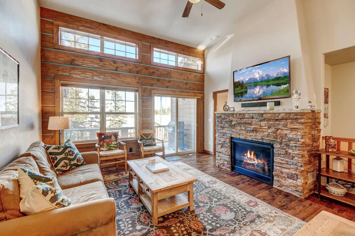 The image showcases a luxurious living room with a glowing fireplace as the centerpiece, surrounded by a plush sectional sofa and a stylish coffee table. Floor-to-ceiling windows flood the space with natural light and offer scenic views, while the setting invites cozy gatherings, relaxation, and enjoying entertainment on the large TV.