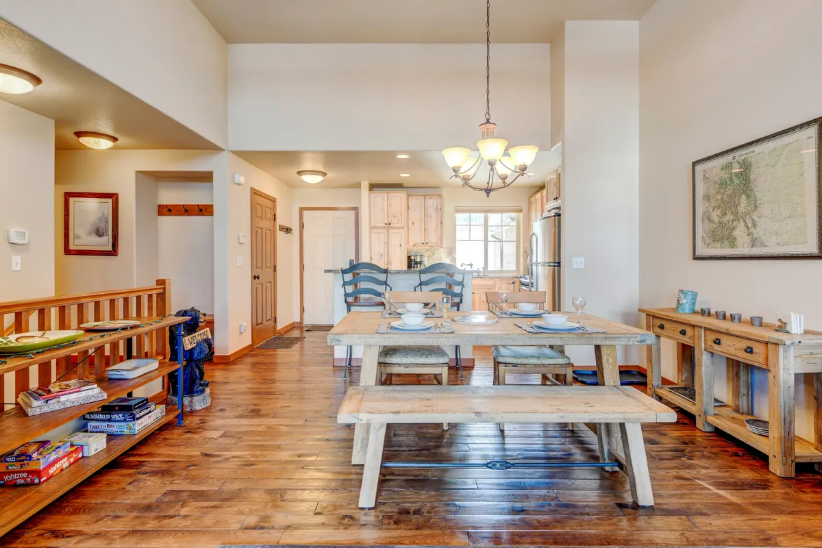 The spacious dining area features a generous table beneath a chandelier, with the kitchen area and wooden shelves visible in the background. The setting is perfect for family meals, planning adventures, or celebrating special occasions.