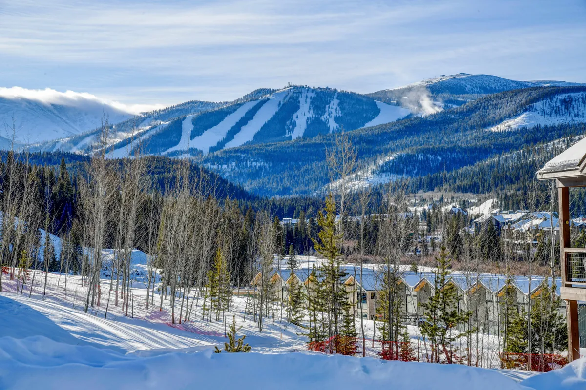 A sweeping view of snow-capped mountains with ski slopes winding down through pine trees. Rows of mountain cabins nestle at the base, and the natural landscape suggests year-round exploration—perfect for skiing, snowboarding, hiking, or photography. The sunlight creates a tranquil yet adventurous mood.