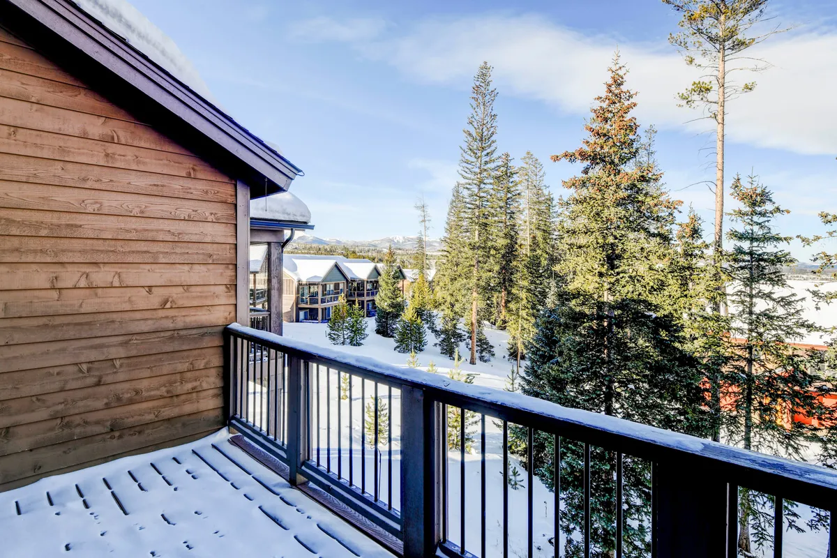 A serene snow-covered balcony overlooks tall evergreen trees and a majestic mountain range in the distance. The outdoor space invites guests to relax and take in the stunning natural surroundings, hinting at the tranquil luxury and mountain views waiting just outside the home.