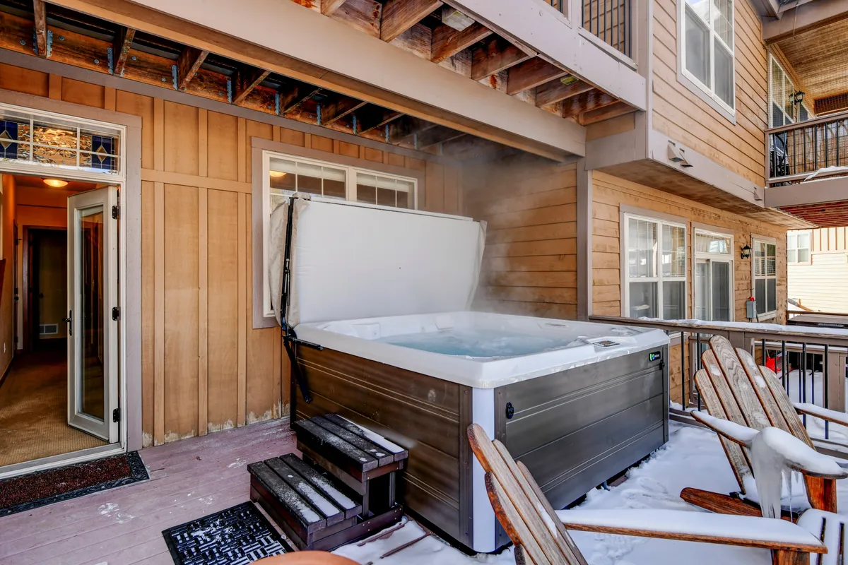 An outdoor area featuring a hot tub on a wooden deck next to the home’s entrance door, with snow on the ground and the promise of sunset views. The space radiates relaxation and social atmosphere.